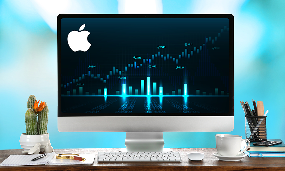 Investing, buying Apple stocks image. An Apple computer placed on a wooden table with a cup of coffee, notes, pens, and a cactus plant. Behind them is a turquoise background.
