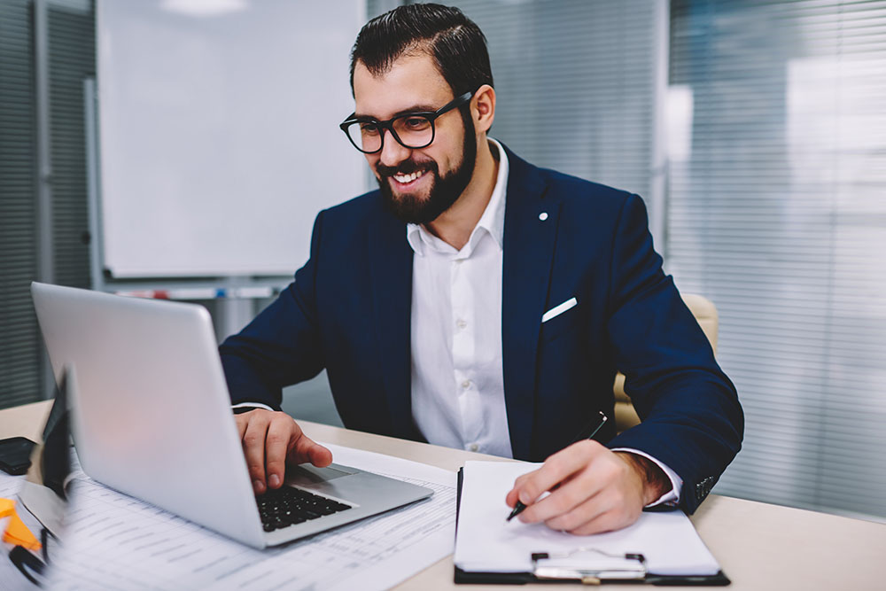 Online trading strategy image. A guy with eye-glasses smiling, wearing a a black suit with white polo holding a pen and using a laptop to create a trading plan or strategy.