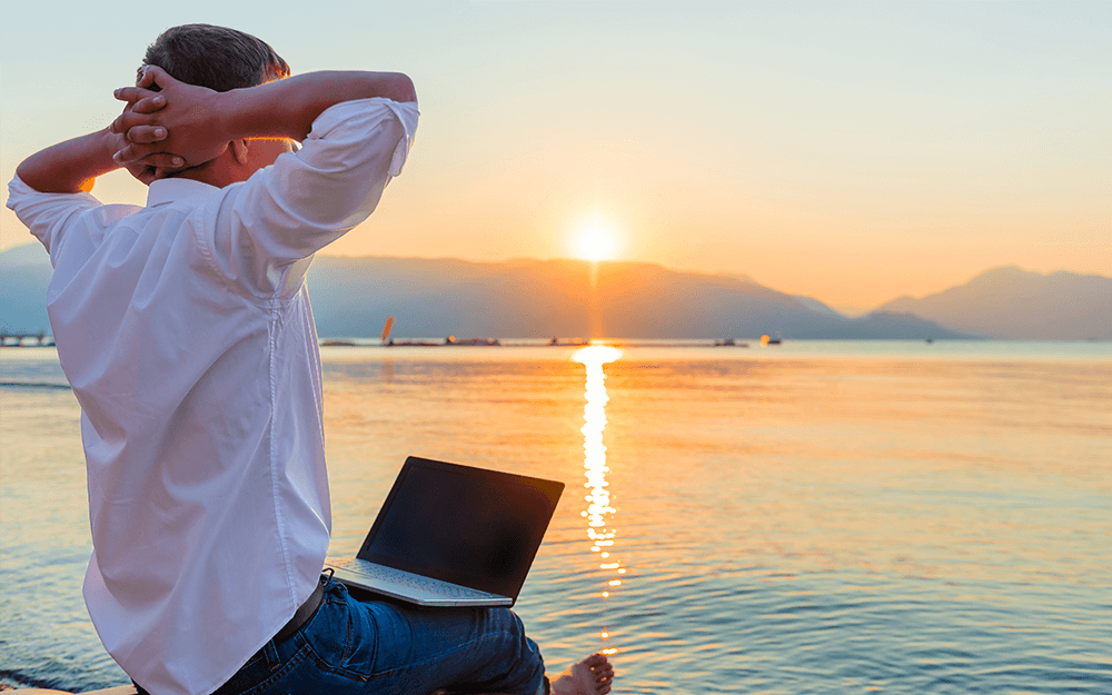 A man working on his laptop while watching the sunset by the beach.